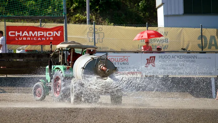 Ein selbstgebauter Motoball-Traktor fährt auf einer staubigen Strecke, während Wasser aus einem Rohr an der Seite spritzt. Im Hintergrund sind Werbebanner und Zuschauer zu sehen.
