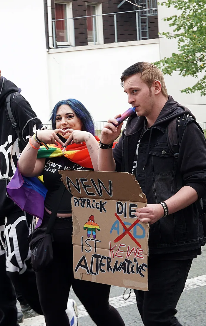 Eine Person mit blauen Haaren trägt eine Regenbogenfahne, während eine andere Person ein Pappschild mit der Aufschrift 'DIE SPD IST KEINE ALTERNATIVE' hält.