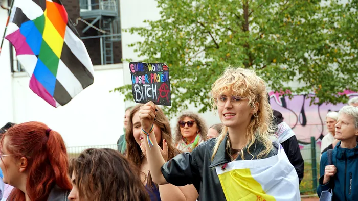 Eine Person mit blonden Locken hält beim Christopher Street Day in Lüdenscheid ein Schild mit der Aufschrift „FEMALE WOMAN UNSER PROJEKT BITCH“ hoch.