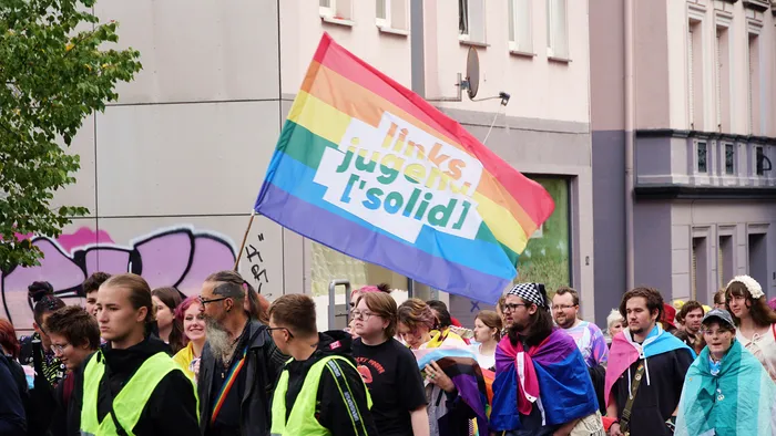 Eine große Regenbogenfahne mit dem weißen Schriftzug „links jugend [solid]“ über einer Menschenmenge bei einer Demonstration.