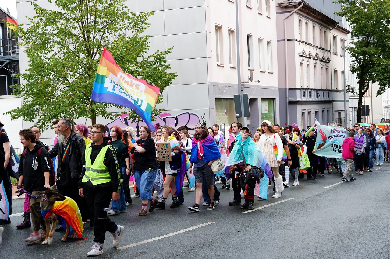 Menschen in bunter Kleidung und mit Regenbogenflaggen ziehen bei einer Parade durch eine Stadtstraße; eine Person hält ein Schild mit der Aufschrift 'LOVE IS NOT HATE'.