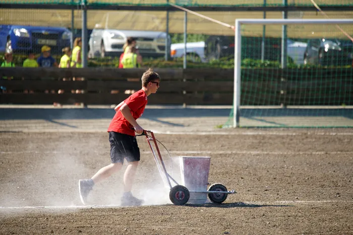 Ein Jugendlicher zieht eine Maschine, die weißen Kalk auf einen Fußballplatz streut. Im Hintergrund stehen Zuschauer und ein Fußballtor.