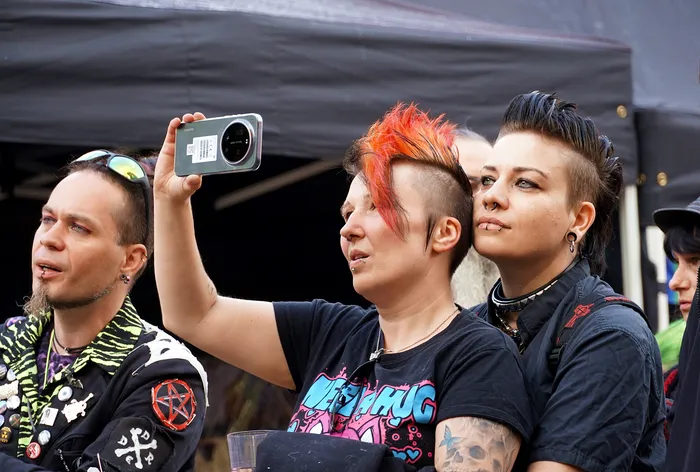 Eine Person mit orangefarbenem Irokesenschnitt macht ein Selfie mit einem Smartphone, flankiert von zwei weiteren Personen im Punk-Stil.