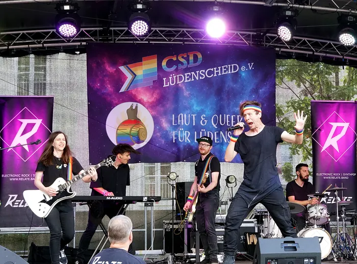Eine Band performt auf einer Bühne vor einem großen Banner mit der Aufschrift „CSD LÜDENSCHEID e.V.“ und Regenbogenmotiven.