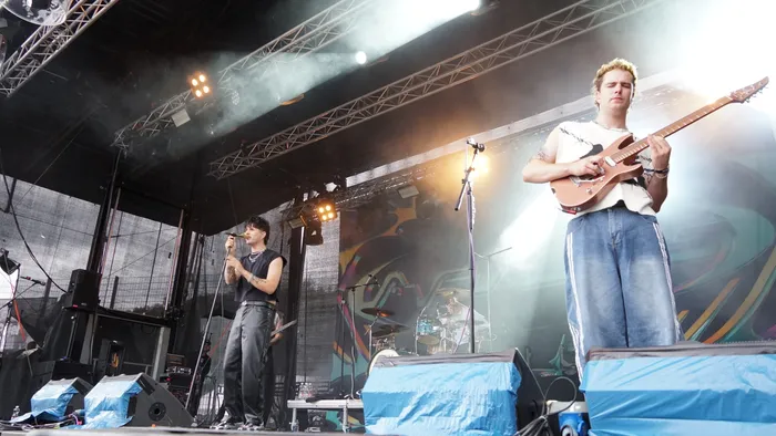Ein Musiker spielt eine E-Gitarre auf einer hell erleuchteten Bühne, während im Hintergrund eine Person singt.