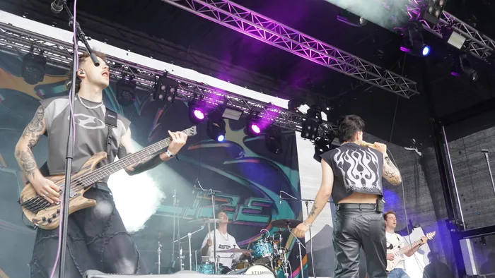 Ein Bassist und eine Person mit gemustertem Oberteil stehen im Vordergrund einer Bühne mit lila Licht und Nebel während eines Live-Auftritts.