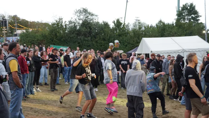 Eine Menschenmenge versammelt sich bei einem Open-Air-Event vor einem weißen Zelt und Bäumen.