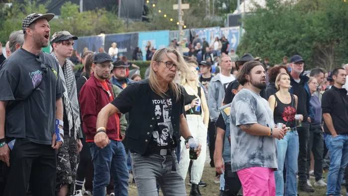 Menschenmenge bei einem Open-Air-Event, im Vordergrund ein Mann mit langen Haaren, Brille und schwarzer Weste, der einen Becher hält.