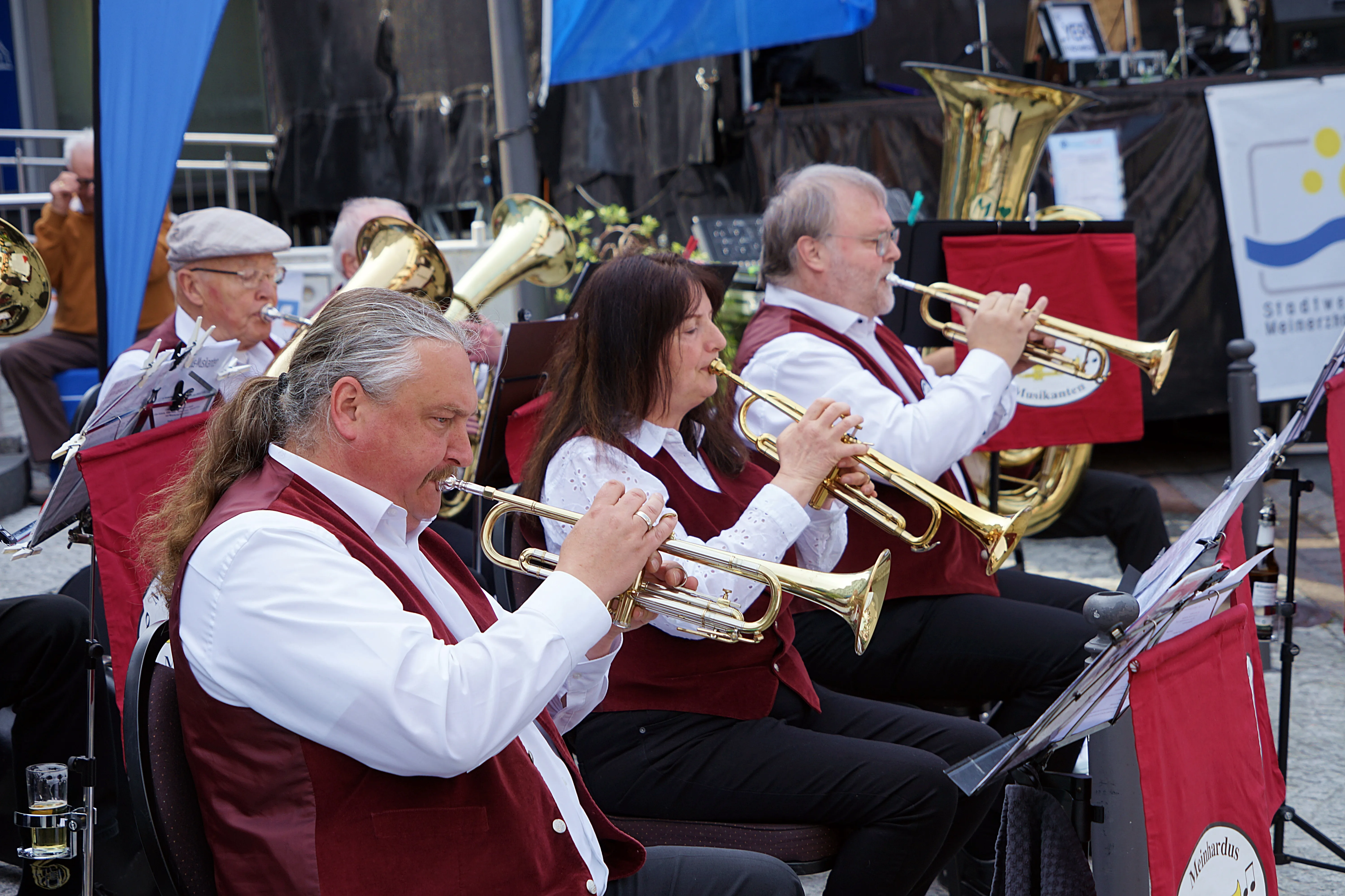 Mehrere Musiker spielen Trompeten im Freien, vor einem Banner mit dem Stadtwappen Meinerzhagens. Im Hintergrund sind weitere Blasinstrumente und ein blaues Zelt zu sehen.