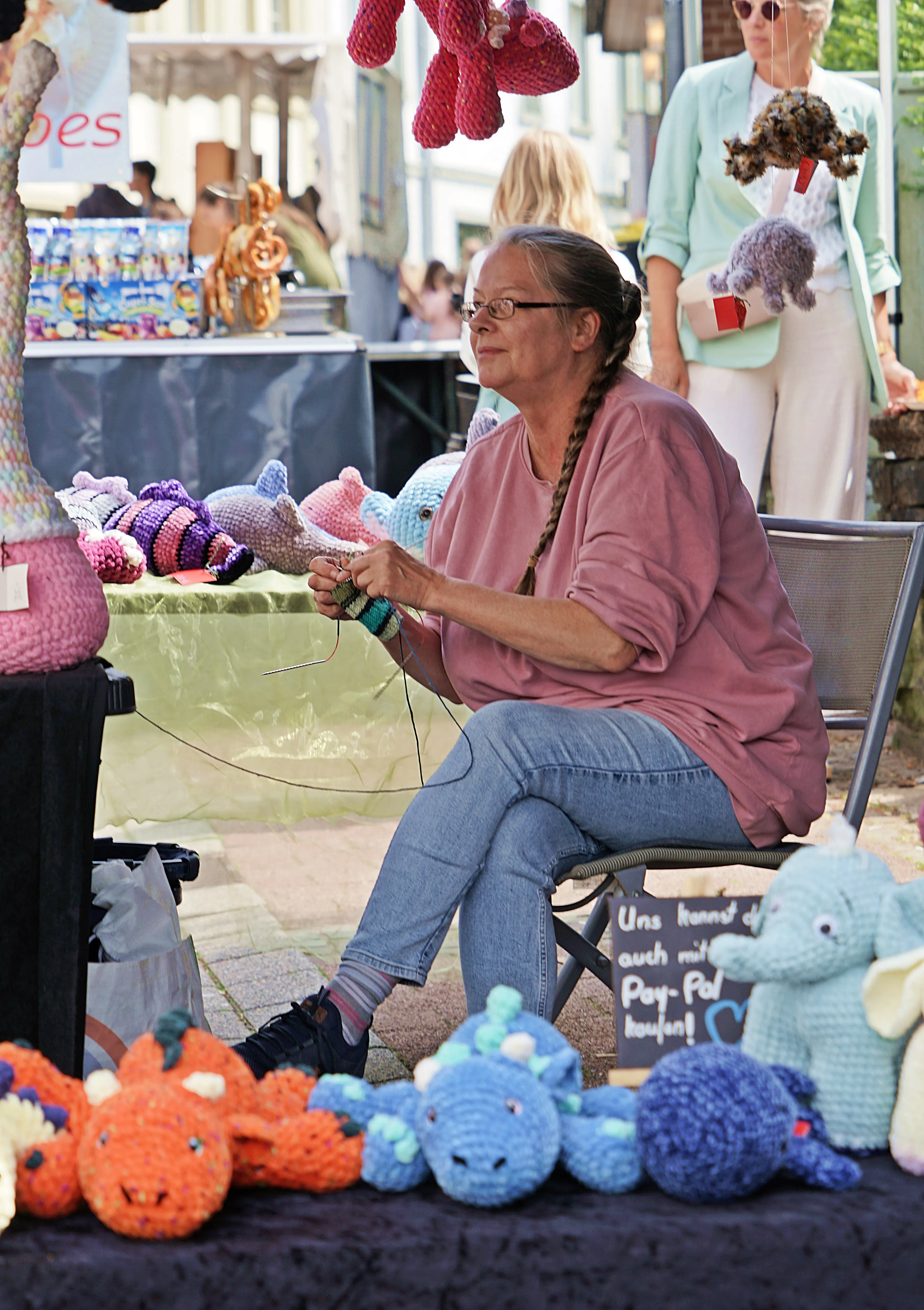 Eine Frau sitzt auf einem Stuhl und strickt bunte Stofftiere. Vor ihr stehen gehäkelte Figuren, im Hintergrund ein Marktstand mit weiteren Handarbeiten.