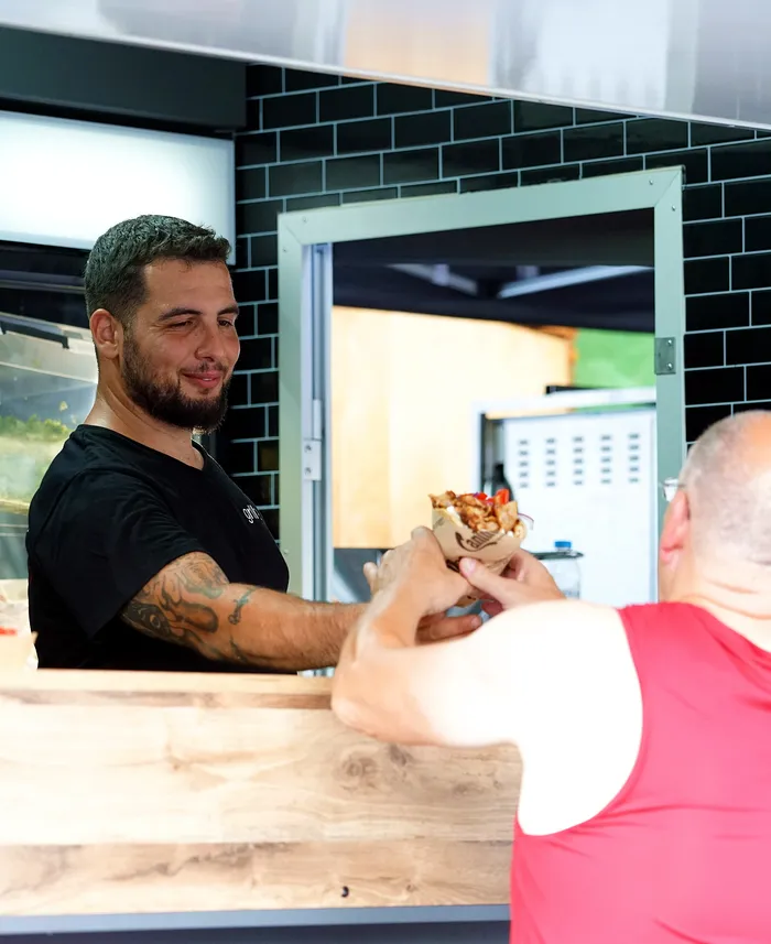 Ein Mann mit Tattoos übergibt einem Kunden in einem Imbiss einen Döner in Alufolie.