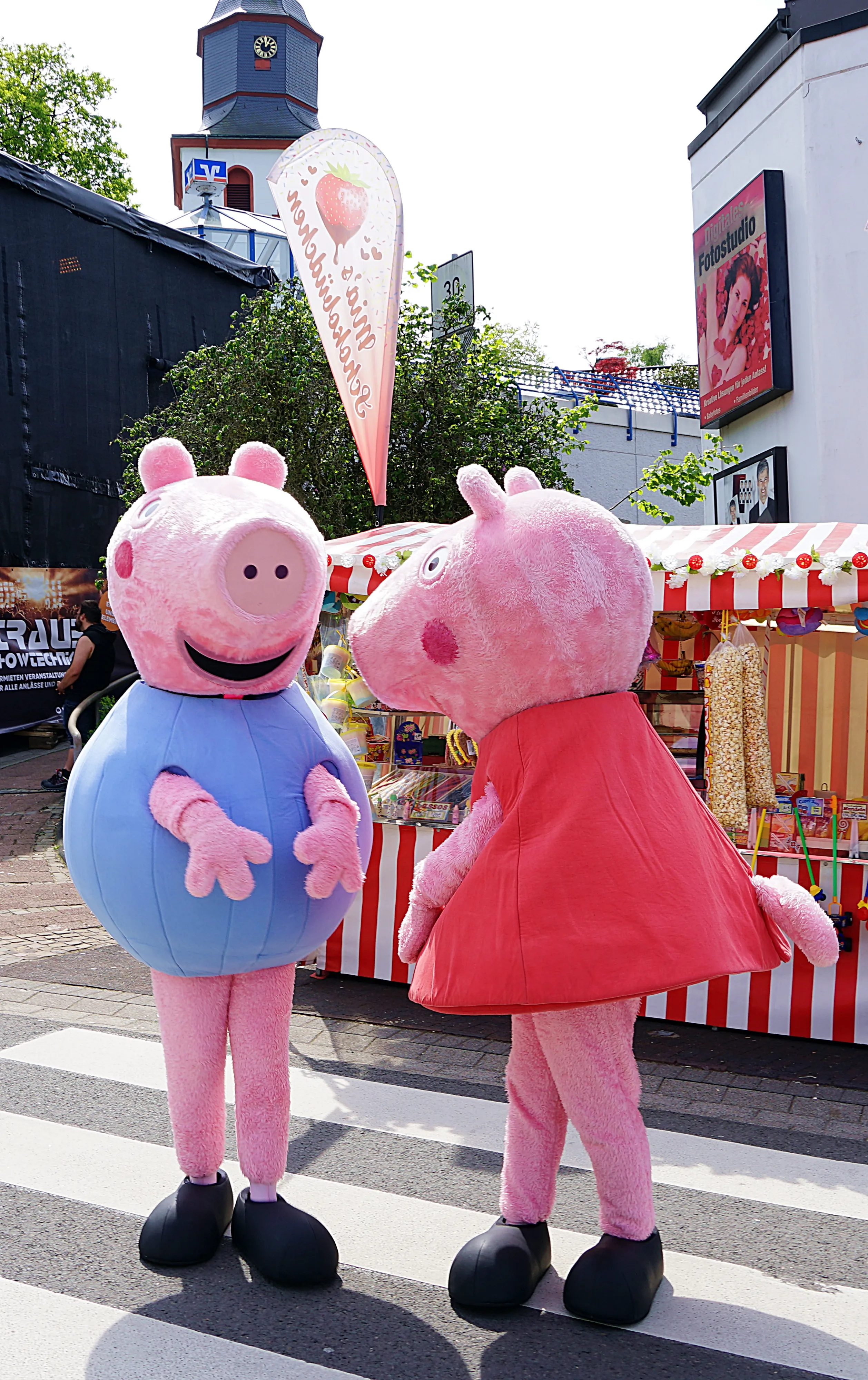 Zwei Personen in Peppa-Wutz-Kostümen stehen auf einer Straße vor einem Marktstand mit Süßigkeiten und einem Eisstand mit Erdbeereis.