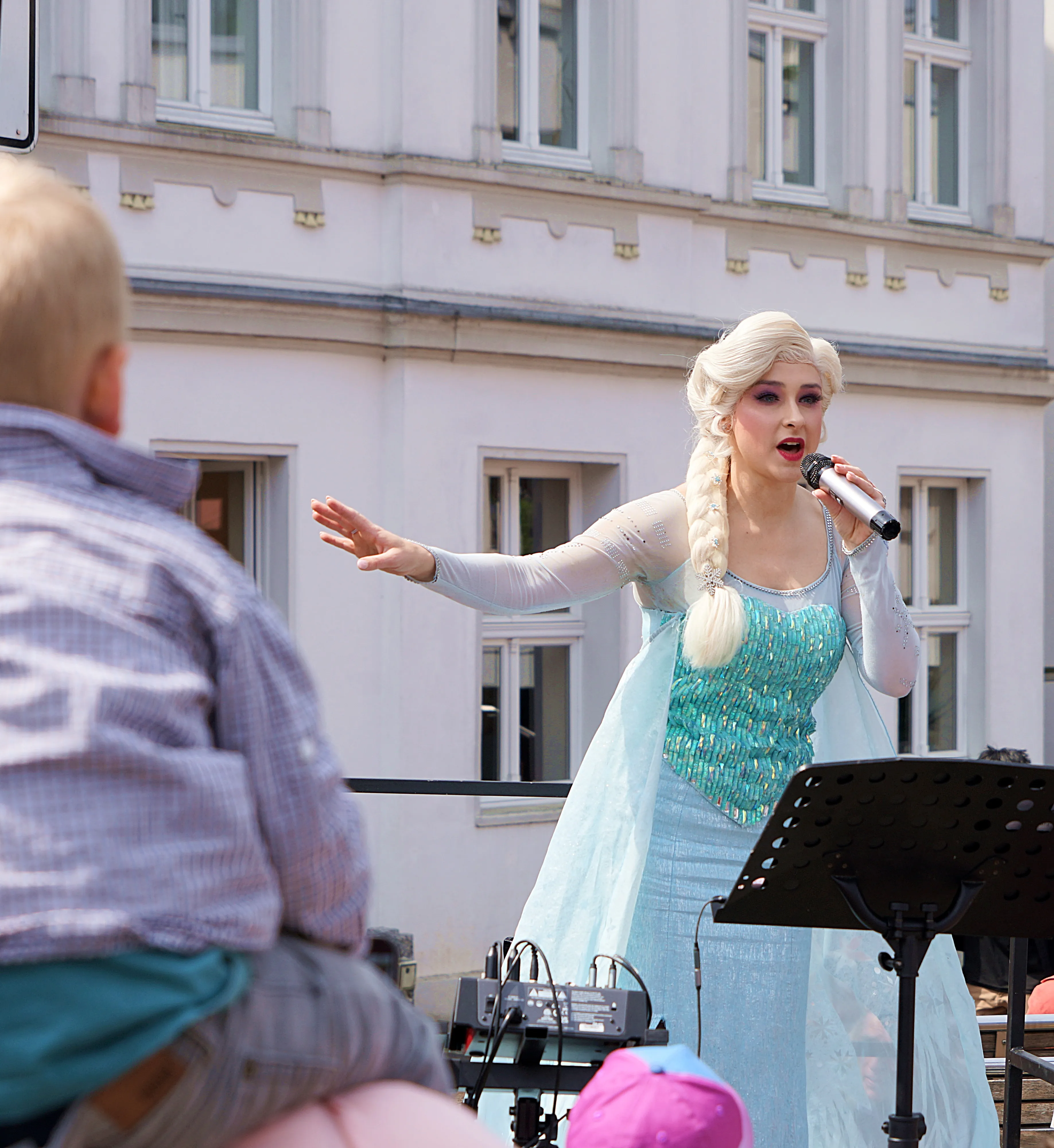 Eine Frau im Kostüm der Eiskönigin Elsa singt mit einem Mikrofon in die Menge. Im Hintergrund sind Gebäude und Zuschauer zu sehen.