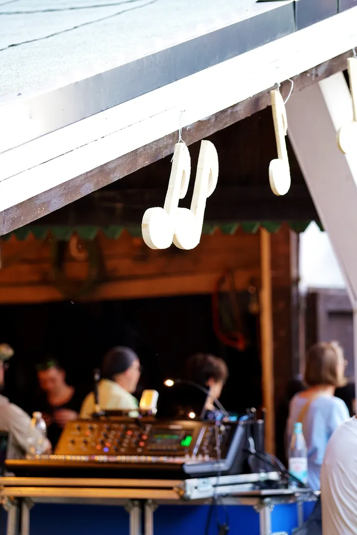 Hanging from the roof of a stage are two large, wooden musical notes. Below, a DJ operates a mixing console with several people visible in the background.