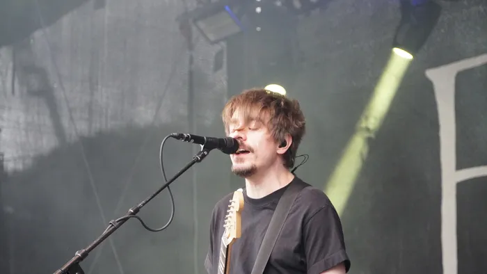 Ein Musiker singt mit geschlossenen Augen in ein Mikrofon und spielt dabei E-Gitarre unter Bühnenlicht.