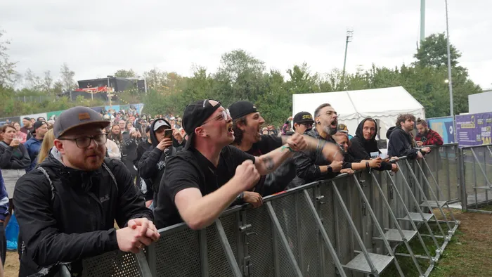 Begeisterte Menschen lehnen an einer Metallabsperrung bei einem Musikfestival und blicken in Richtung der Bühne.