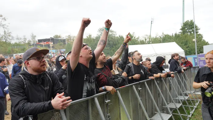 Menschen jubeln mit erhobenen Armen und Fäusten hinter einer Metallabsperrung bei einem Musikfestival.