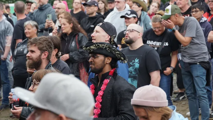 Ein Mann mit Sonnenbrille, einem verzierten schwarzen Hut und einer rosa Blumenkette steht in einer Menschenmenge.