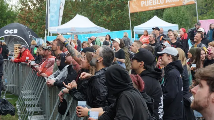 Eine Menschenmenge steht hinter einer Metallabsperrung bei einem Musikfestival und blickt in Richtung der Bühne.