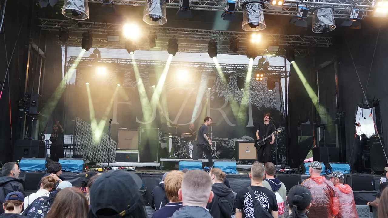 Die Band Fjort spielt auf einer hell erleuchteten Bühne vor einem Publikum. Im Hintergrund ist ein großes Banner mit dem Schriftzug „FJORT“ zu sehen.