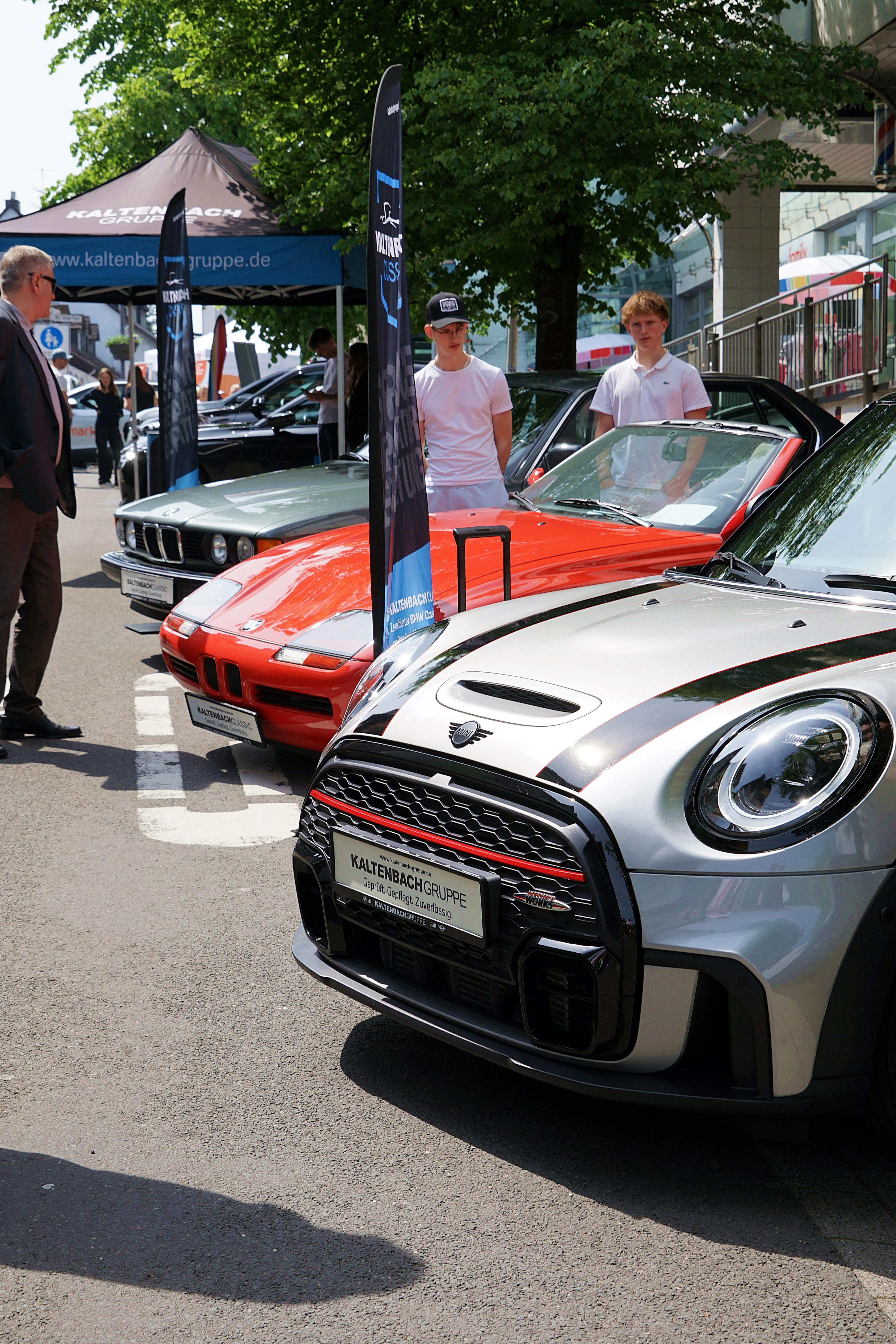 Silberner Aston Martin steht im Vordergrund einer Oldtimer-Ausstellung mit weiteren Fahrzeugen und Besuchern im Hintergrund.