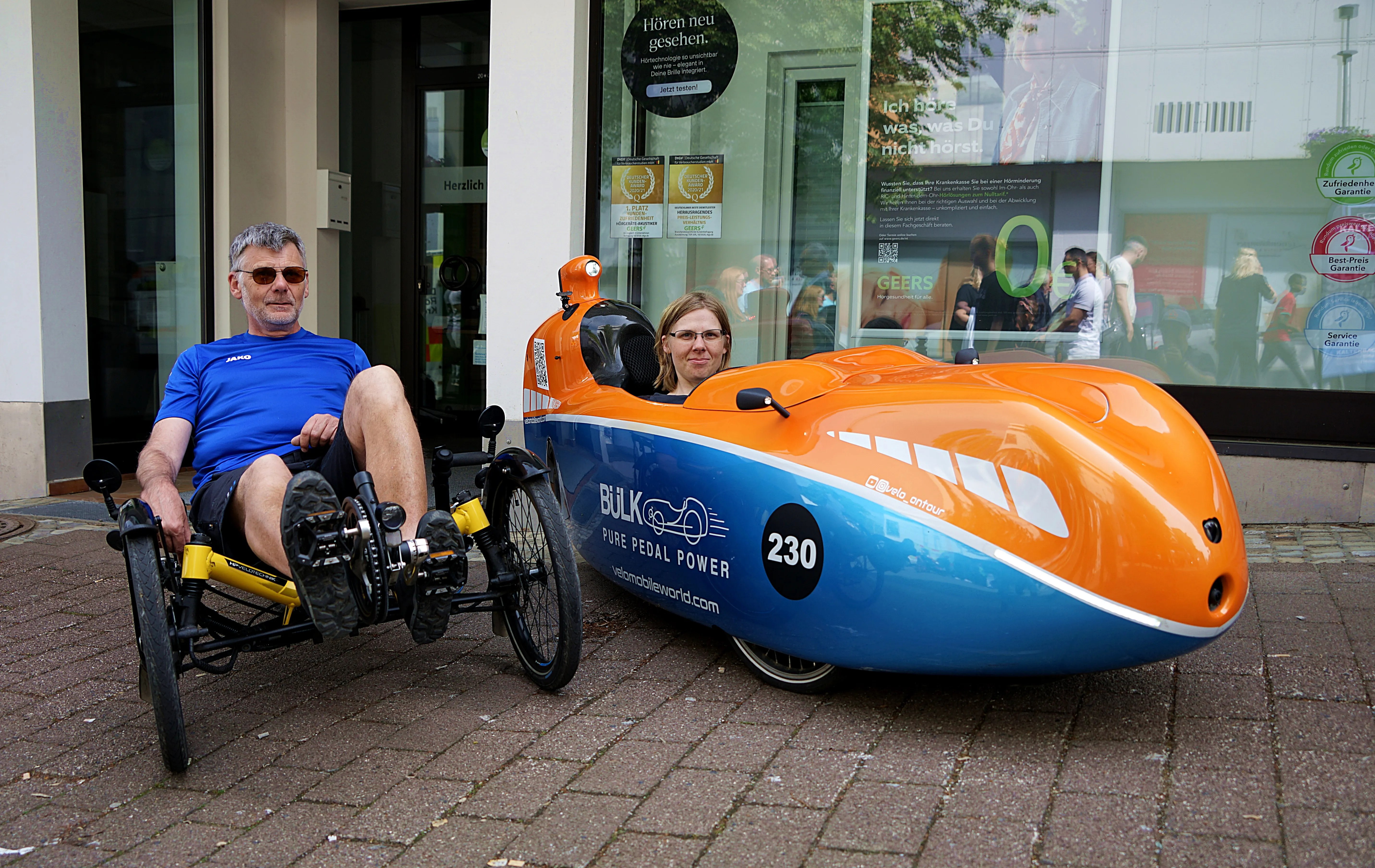 Ein Mann sitzt in einem blauen Liegedreirad, während eine Frau in einem orange-blauen, aerodynamischen Fahrzeug sitzt. Das Fahrzeug trägt die Aufschrift „Bükl – Pure Pedal Power“ und die Nummer 230.