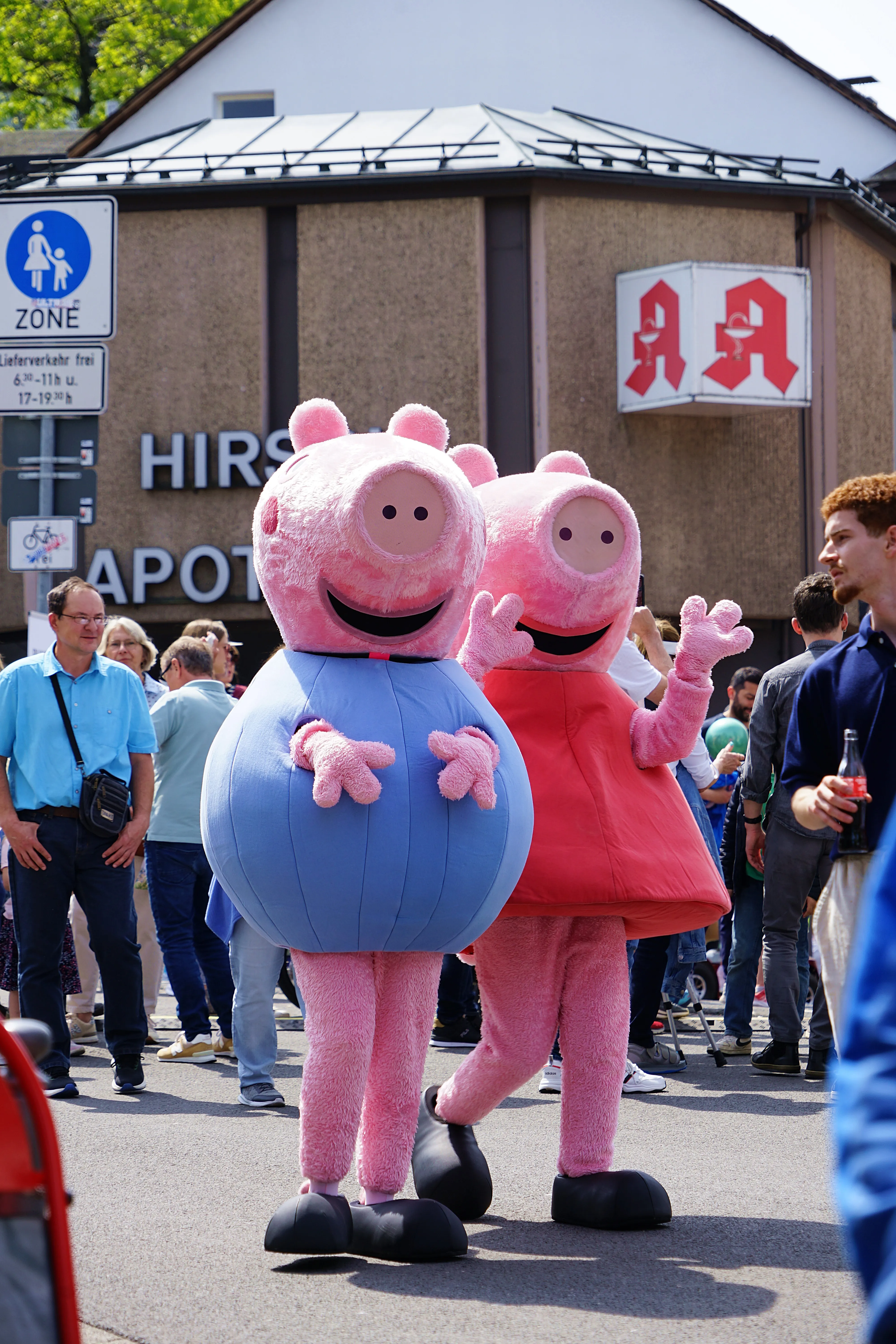 Zwei Personen in Peppa-Wutz-Kostümen, eine in Rosa und eine in Hellblau, stehen auf einer Straße vor Passanten und Geschäften.