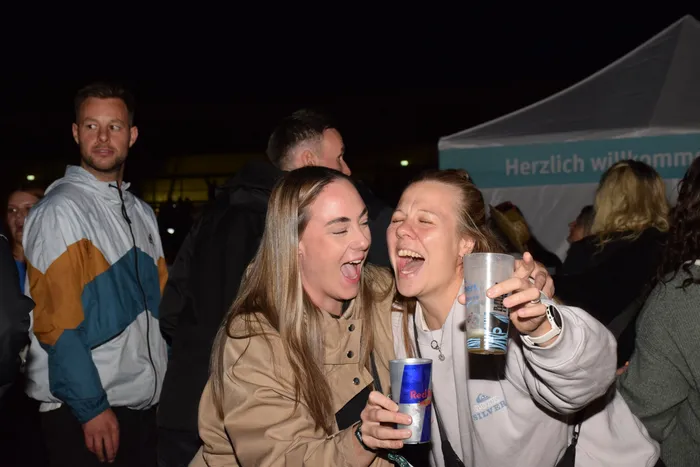 Zwei lachende Frauen feiern nachts bei einem Event; eine hält eine Red Bull Dose, die andere einen Plastikbecher.