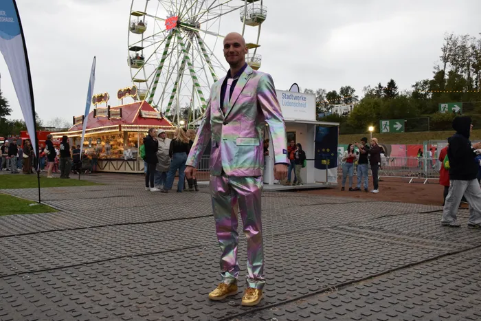 Ein Mann in einem schillernden, holografischen Anzug und goldenen Schuhen steht auf einem Festplatz vor einem Riesenrad.