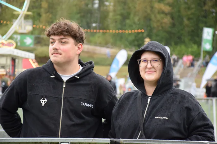 Ein junger Mann und eine junge Frau in schwarzen Kapuzenpullovern lächeln auf einem Festgelände im Regen.
