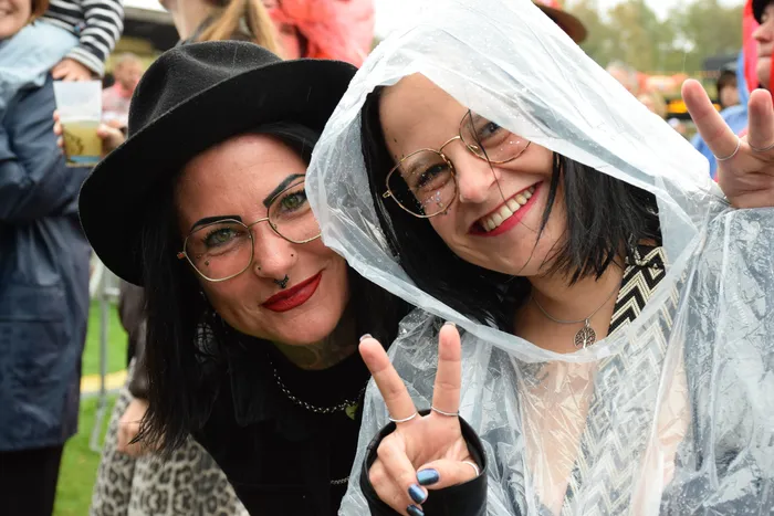 Zwei lächelnde Frauen mit Brillen zeigen Peace-Zeichen, eine trägt einen schwarzen Hut und die andere einen durchsichtigen Regenmantel.