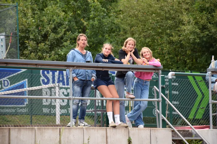 Vier junge Frauen lehnen an einem Metallgeländer auf einer Tennisanlage.