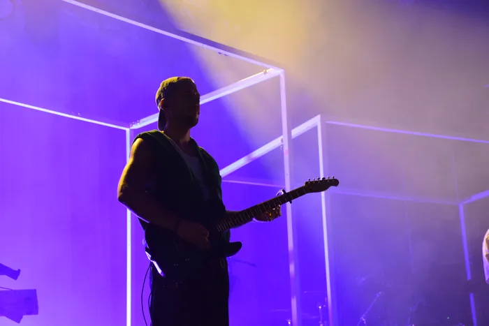 Ein Musiker der Band ClockClock spielt eine E-Gitarre unter violettem und gelbem Bühnenlicht.