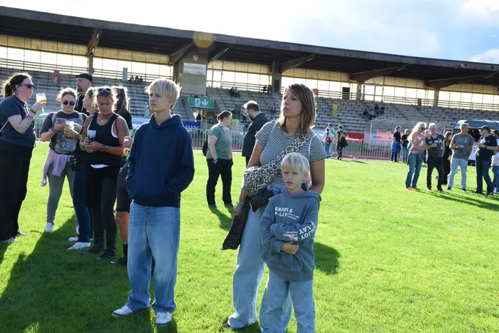 Besucher stehen auf einer Rasenfläche vor einer großen Stadiontribüne bei sonnigem Wetter.