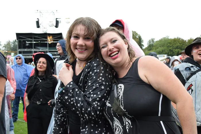 Zwei lachende Frauen lehnen ihre Köpfe bei Regen auf einem Festival aneinander; eine trägt eine Jacke mit Sternenmuster.