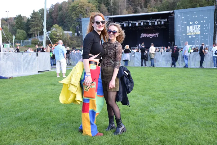 Zwei lächelnde Frauen posieren vor einer Festivalbühne beim Baltz Festival; eine trägt bunte Patchwork-Hosen, die andere ein Leopardenmuster-Oberteil.
