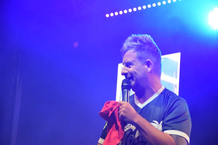 Almklausi singt unter blauem Bühnenlicht in ein Mikrofon und hält ein rotes Tuch.
