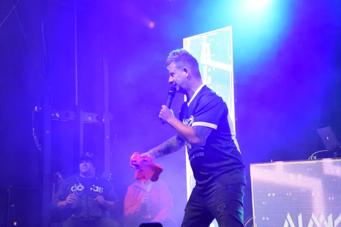 Almklausi singt unter blauem Bühnenlicht in ein Mikrofon und hält ein rotes Tuch.