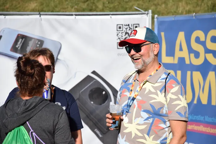 Ein lächelnder Mann mit Sonnenbrille, roter Kappe und tropischem Hemd hält ein Getränk und unterhält sich mit anderen Personen bei der Bautz 2026.