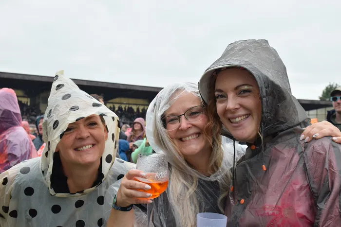 Drei lächelnde Frauen in Regenponchos beim Bautz-Samstag, eine hält ein Glas mit einem orangefarbenen Getränk.