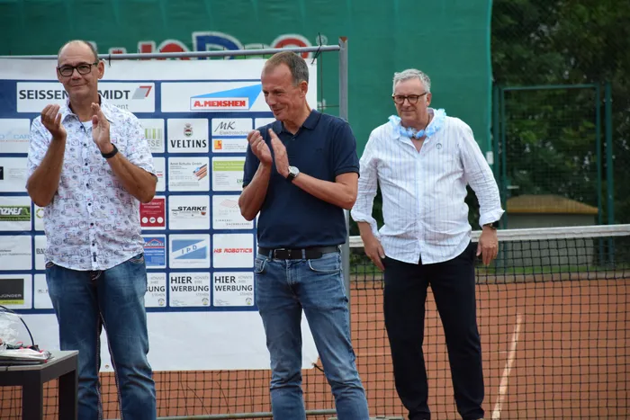 Drei Männer stehen vor einer Sponsorenwand auf einem Tennisplatz während der Jubiläumsfeier der MTC/TC Halver Open.