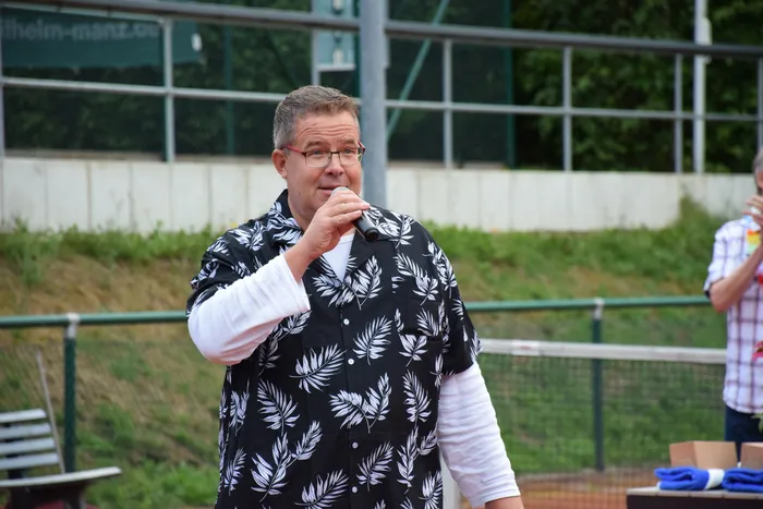 Ein Mann mit Brille und einem schwarz-weiß gemusterten Hemd spricht in ein Mikrofon bei der Jubiläumsfeier der MTC/TC Halver Open auf einer Tennisanlage.