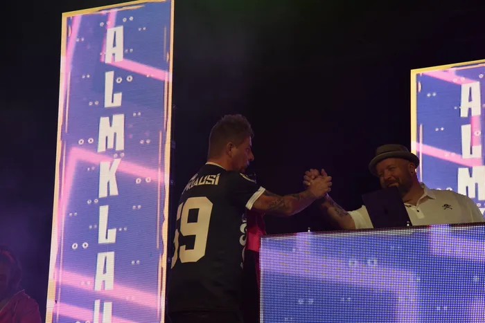 Almklausi in einem dunklen Trikot schüttelt einem Mann mit Hut hinter einem DJ-Pult die Hand vor großen LED-Wänden.