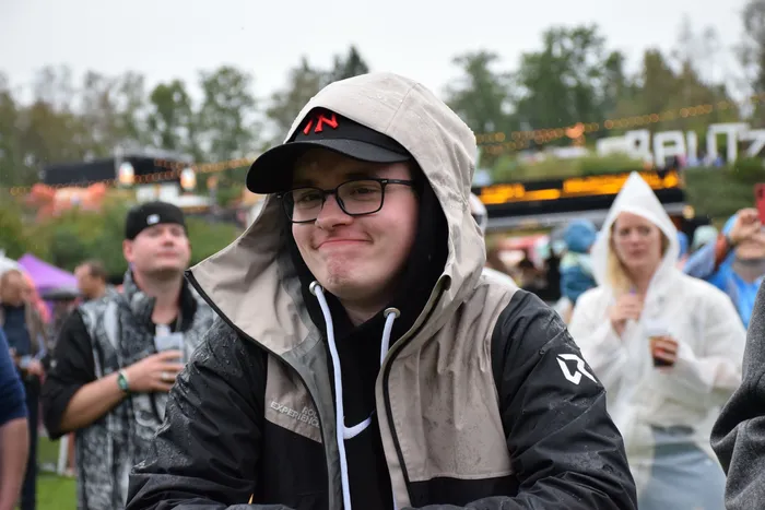 Ein lächelnder junger Mann mit Brille und Kapuzenjacke bei einer Veranstaltung im Freien im Regen.