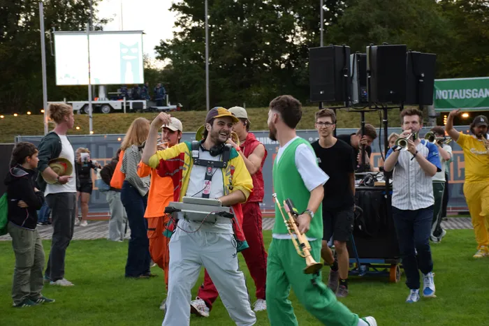 Musiker in bunten Outfits spielen Blasinstrumente inmitten eines Publikums bei einer Open-Air-Veranstaltung.