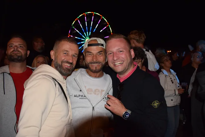 Vier lächelnde Männer stehen bei einer nächtlichen Veranstaltung vor einem bunt beleuchteten Riesenrad.