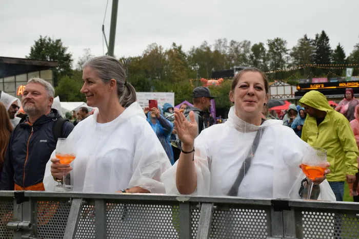 Zwei Frauen in weißen Regenponchos stehen hinter einer Metallabsperrung beim Bautz-Samstag; eine hält ein Glas mit einem orangefarbenen Getränk.