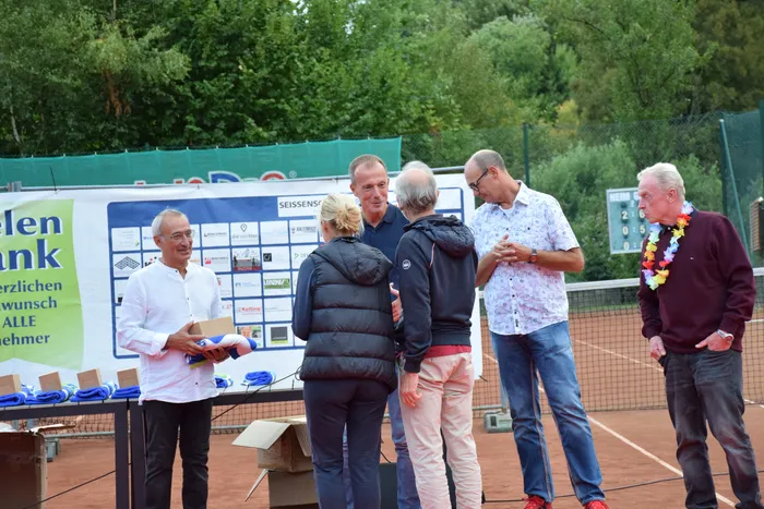 Männer bei der Jubiläumsfeier der MTC/TC Halver Open auf einem Tennisplatz; ein Mann in weißem Hemd hält eine Box und ein blaues Tuch.