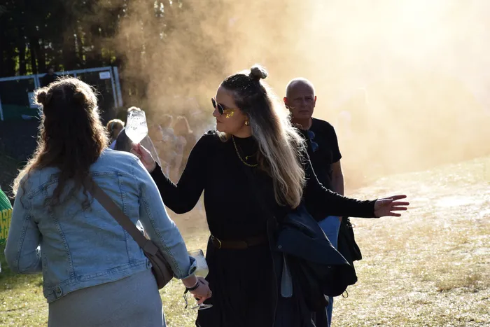 Eine Frau in einem schwarzen Kleid und Sonnenbrille steht mit ausgebreiteten Armen in einer sonnendurchfluteten, staubigen Umgebung.