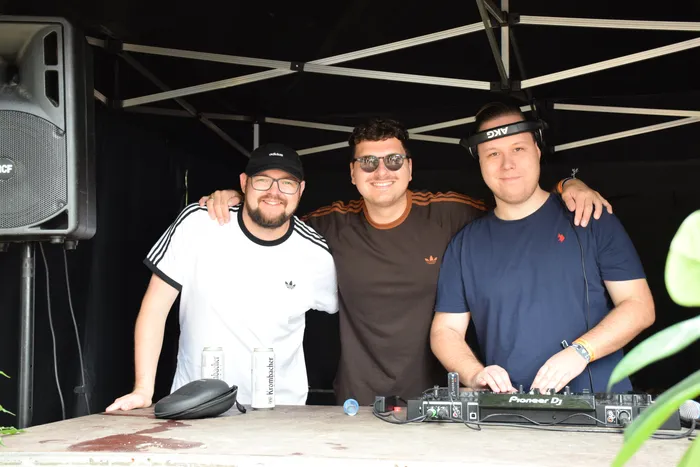 Drei lächelnde Männer stehen hinter einem Tisch mit DJ-Equipment und Krombacher-Dosen unter einem schwarzen Zeltdach.